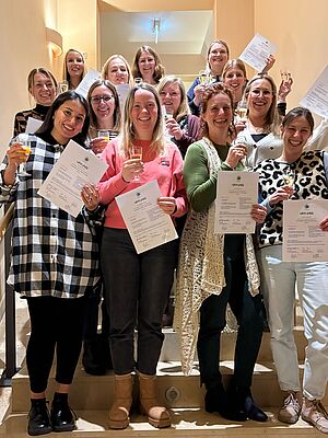 Eine Gruppe von Frauen steht fröhlich auf einer Treppe in einem Innenraum. Sie halten Zertifikate und Gläser in der Hand und lächeln stolz in die Kamera. Die Szene wirkt festlich und zeigt den Abschluss einer gelungenen Ausbildung oder Veranstaltung.