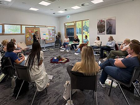 „Teilnehmerinnen einer Ausbildung zur Eltern-Kind-Gruppenleiterin sitzen in einem Stuhlkreis in einem Seminarraum. Vorne erklärt eine Frau etwas an einem Flipchart, im Hintergrund sind Pinnwände mit bunten Moderationskarten zu sehen.“