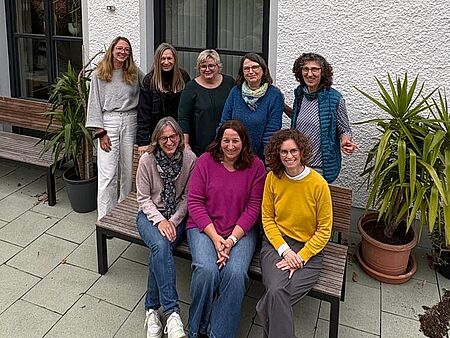 Vor dem Tagungshotel „Der Hermannsberg“ in Wiesent sitzt und steht ein Teil des EKG-Fortbildungsteams für ein Gruppenfoto zusammen. Acht Frauen haben sich lächelnd vor der weißen Hausfassade mit dem bunten „H“-Logo versammelt. Die entspannte Atmosphäre spiegelt die Freude am gemeinsamen Arbeiten und Austauschen wider.