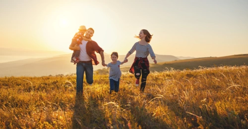 Eine vierköpfige Familie läuft lachend über eine Wiese in der Abendsonne. Der Vater trägt ein kleines Kind auf dem Arm und hält die Hand eines älteren Kindes. Die Mutter läuft daneben und hält ebenfalls die Hand des älteren Kindes. Alle sind fröhlich, die Sonne steht tief am Horizont und wirft warmes Licht auf die hügelige Landschaft.