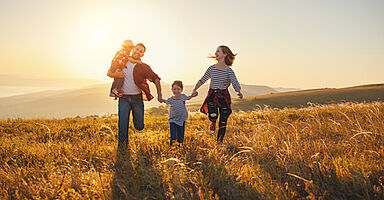 Eine vierköpfige Familie läuft lachend über eine Wiese in der Abendsonne. Der Vater trägt ein kleines Kind auf dem Arm und hält die Hand eines älteren Kindes. Die Mutter läuft daneben und hält ebenfalls die Hand des älteren Kindes. Alle sind fröhlich, die Sonne steht tief am Horizont und wirft warmes Licht auf die hügelige Landschaft.