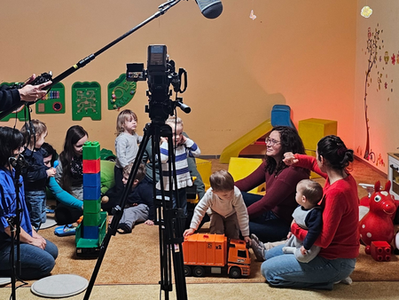 In einem bunt dekorierten Spielraum sitzen mehrere Kleinkinder und Erwachsene auf dem Boden und spielen mit Bauklötzen und Spielzeug, während ein Kamerateam mit Kamera auf Stativ und Tonangel einen Werbespot filmt. Im Vordergrund steht eine Filmkamera, darüber hängt ein Mikrofon.