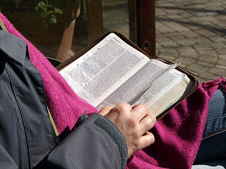 Eine Person mit Jacke und pinkfarbenem Schal sitzt auf einem Stuhl und blättert in einer Bibel.