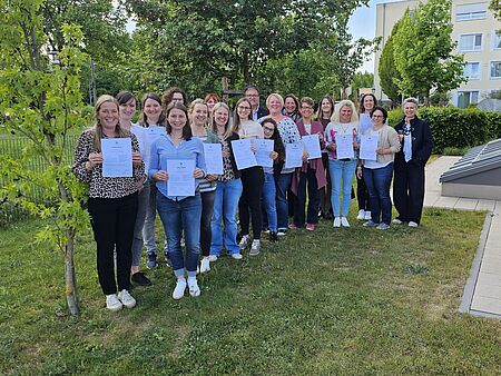 „Gruppenfoto von etwa 18 Erwachsenen im Freien auf einer Grünfläche zwischen Gebäuden. Die Personen stehen in mehreren Reihen und halten Urkunden oder Zertifikate in die Kamera. Im Hintergrund sind Bäume, Büsche und Wohngebäude zu sehen. Die Stimmung wirkt freundlich und feierlich.“