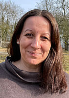 Eine Frau mit langen, dunklen Haaren steht im Freien und lächelt in die Kamera. Sie trägt einen braunen Pullover mit weitem Kragen. Im Hintergrund sind Bäume und eine Wiese bei sonnigem Wetter zu sehen.