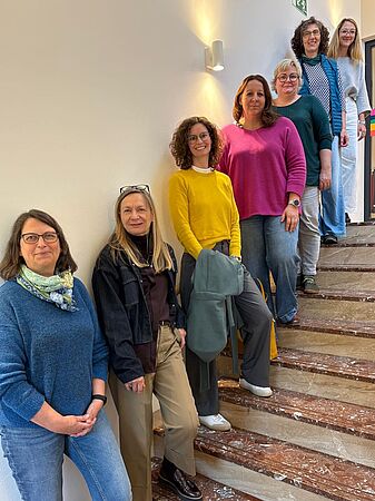 Acht Frauen stehen lächelnd auf einer Treppe in einem Gebäude und blicken zur Kamera.