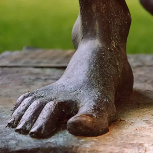 Die nackten Füße einer Bronzestatue des Hl. Franz von Assisi berühren den Steinboden. Im Hintergrund ist Licht und grünes Gras zu sehen.