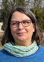 Porträt einer Frau mit dunklem, lockigem Haar, die eine Brille trägt und in die Kamera lächelt. Sie trägt einen blauen Pullover und einen türkisfarbenen, gehäkelten Schal. Im Hintergrund sind Bäume und ein Gebäude bei Tageslicht zu sehen.
