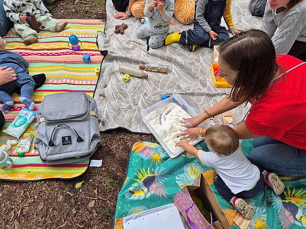 Mehrere kleine Kinder sitzen mit Erwachsenen auf bunten Decken im Freien. Eine Frau hilft einem Kleinkind, mit den Händen in einer Wanne mit weißem Material (wahrscheinlich Mehl oder Sand) zu spielen. Um sie herum liegen Spielzeuge, Trinkflaschen, eine Wickeltasche und andere Babyutensilien. Die Szene wirkt wie ein gemeinsames Outdoor-Spiel- oder Lernangebot für Kleinkinder.
