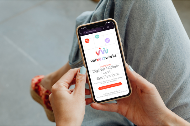 Person hält ein Smartphone mit der Startseite von verNETZwerkt und dem Hinweis „Coming Soon – Digitaler Rückenwind fürs Ehrenamt“.