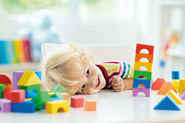 Ein blondes Kleinkind liegt lachend auf einem Tisch und spielt mit bunten Bauklötzen. Das Kind trägt ein gestreiftes T-Shirt in Regenbogenfarben. Um es herum sind viele verschiedenfarbige und geometrisch geformte Holzbausteine verteilt. Die Szene wirkt fröhlich und farbenfroh, mit hellem Tageslicht im Hintergrund.