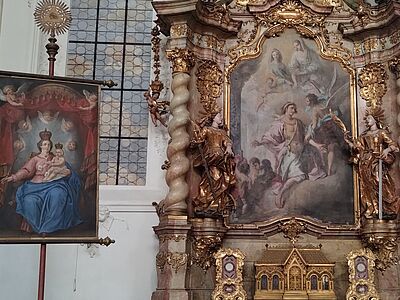 Das Bild zeigt rechts einen Seitenaltar im Stil des Rokoko. Auf dessen Altarbild sind Engel zu sehen. Links davon ist ein Bild der Jungfrau Maria mit Jesuskind zu sehen. Den Hintergrund bildet ein Kirchenfenster.