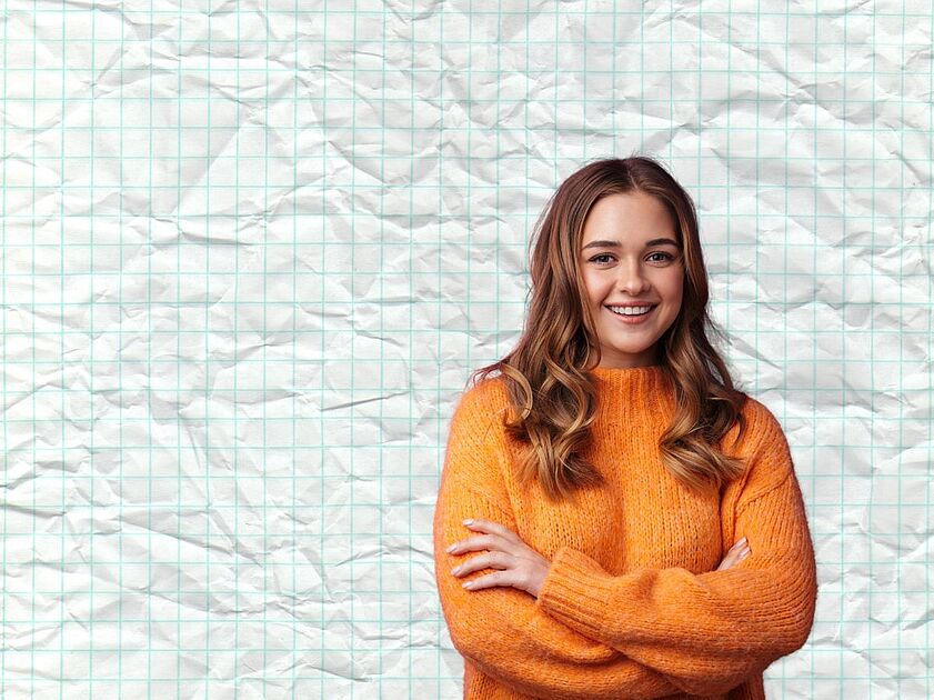 Eine junge frau im orangefarbenen Strickpullover steht mit verschränkten Armen da und blickt zufrieden.