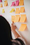 Person mit dunklen Haaren zeigt mit dem Finger auf bunte Haftnotizen an einer weißen Wand, überwiegend orange und gelbe Post-its mit handschriftlichen Notizen, einzelne pinke Zettel dazwischen, typische Brainstorming- oder Workshop-Szene