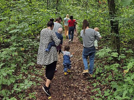 Eine Gruppe von Müttern und kleinen Kindern geht gemeinsam auf einem schmalen Waldweg. Die Erwachsenen tragen zum Teil Babys auf dem Arm, während Kleinkinder neben ihnen laufen. Rundherum ist dichter, grüner Wald – eine Szene, die Gemeinschaft und Naturverbundenheit ausstrahlt.