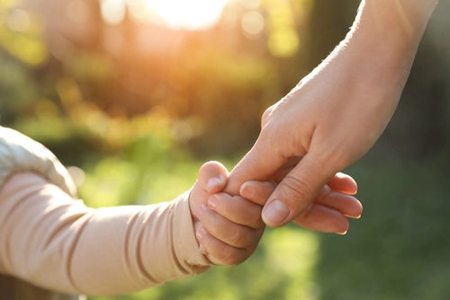 Ein kleines Kind hält die Hand eines Erwachsenen bei Sonnenuntergang im Freien. Die Szene vermittelt Wärme, Geborgenheit und Vertrauen, während weiches Sonnenlicht die Umgebung erhellt.