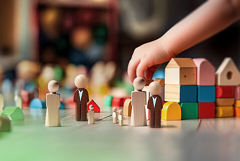 Ein Kind spielt mit Holzfiguren, die Menschen in verschiedenen Größen darstellen, vor einem bunten Bauwerk aus Bauklötzen. Die Szene symbolisiert eine Familie in einem fantasievollen Spielumfeld.