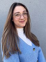 Junge Frau mit langen braunen Haaren, Brille und hellblauem Strickpullover steht vor einer grauen Wand und lächelt leicht in die Kamera.