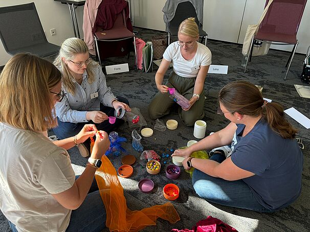 Vier Frauen sitzen in einem Kreis auf dem Boden und beschäftigen sich mit bunten Materialien wie Tüchern, Dosen, Murmeln und kleinen Objekten. Sie wirken konzentriert und arbeiten gemeinsam kreativ. Im Hintergrund stehen Stühle mit Namensschildern und abgelegten Taschen