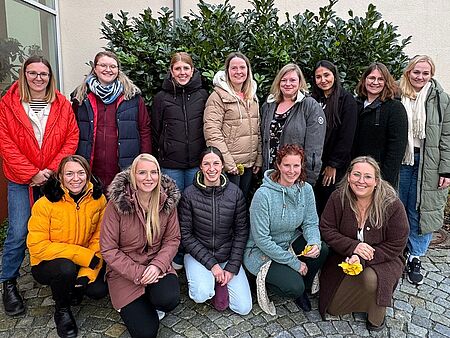 Eine Gruppe von vierzehn Frauen steht und kniet in zwei Reihen im Freien vor einer hellen Hauswand und grünen Sträuchern. Sie tragen winterliche Jacken in verschiedenen Farben, lächeln in die Kamera und wirken fröhlich und verbunden. Einige halten gelbe Papierblätter in den Händen.